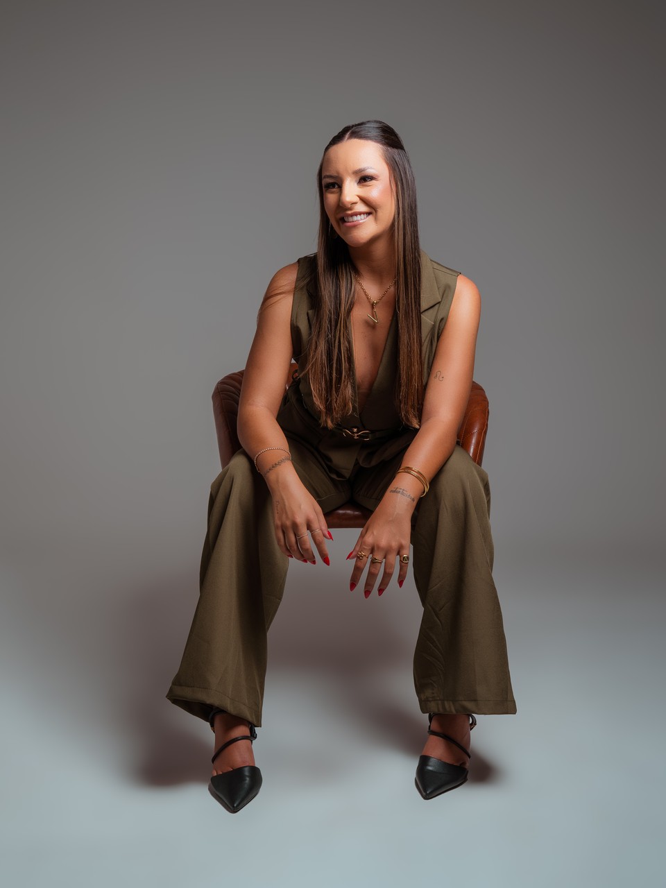 Vitória sentada em uma poltrona de couro, rindo, de conjunto de alfaiataria verde militar.