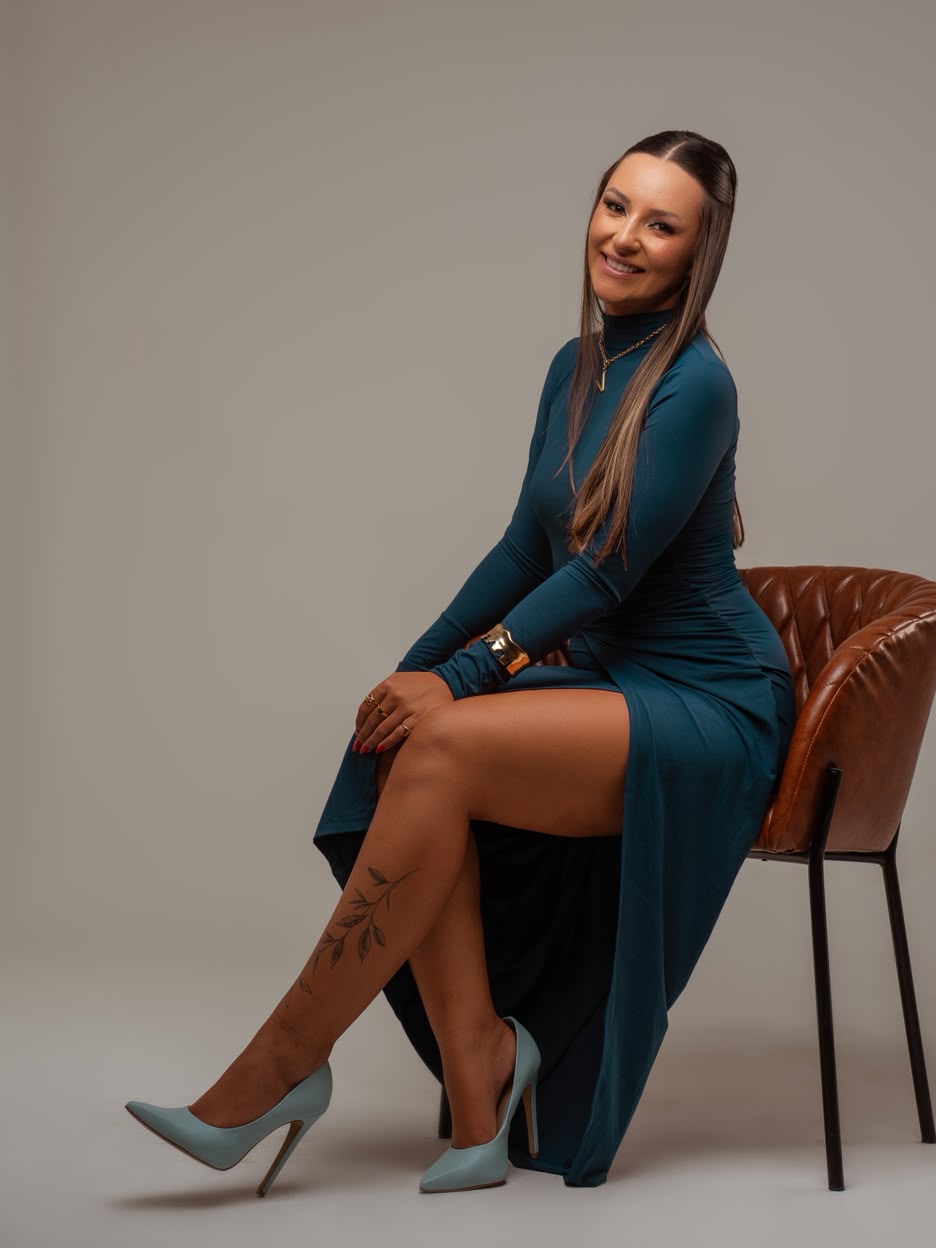 Vitória sentada em uma poltrona caramelo, de vestido azul petróleo de manga longa.